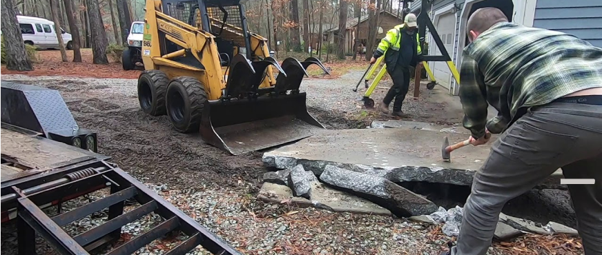 A yellow skid steer loader with a grapple attachment assisting in the manual breaking and removal of a concrete slab on a residential site