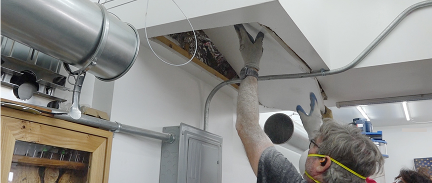 Worker in safety gear accessing overhead ceiling area with exposed insulation, wiring, and ductwork during professional ceiling grid and HVAC duct removal services in New Jersey.