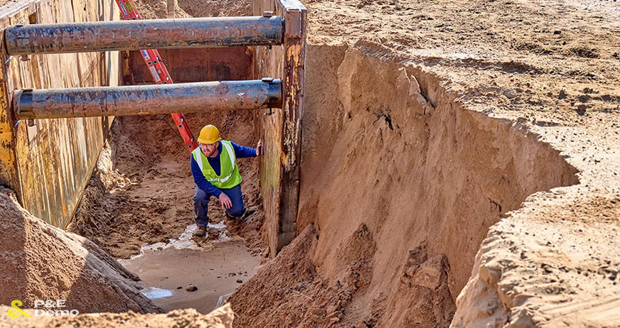 A worker in a safety vest and hard hat inspecting a deep utility trench reinforced with steel shoring boxes by P&E Demolition.