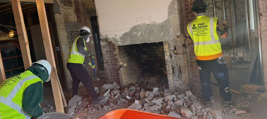 NJ demolition crew dismantling a brick fireplace indoors, wearing safety gear and high‑visibility vests, during a professional hearth removal project.