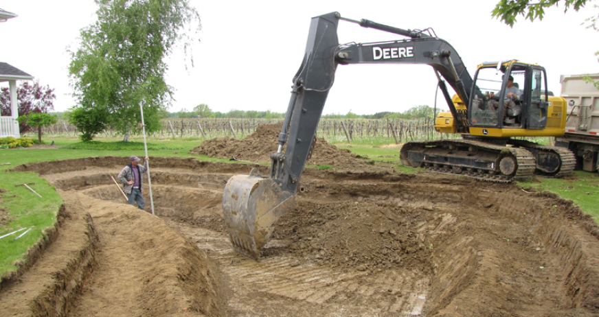 Excavator digging out a decorative pond while a worker measures depth during professional pond removal services in New Jersey