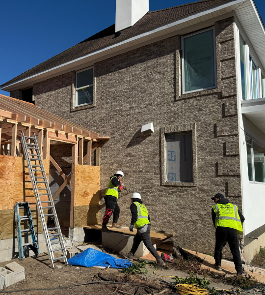 Four construction workers in safety vests and helmets working beside a brick house with tools and materials around them.