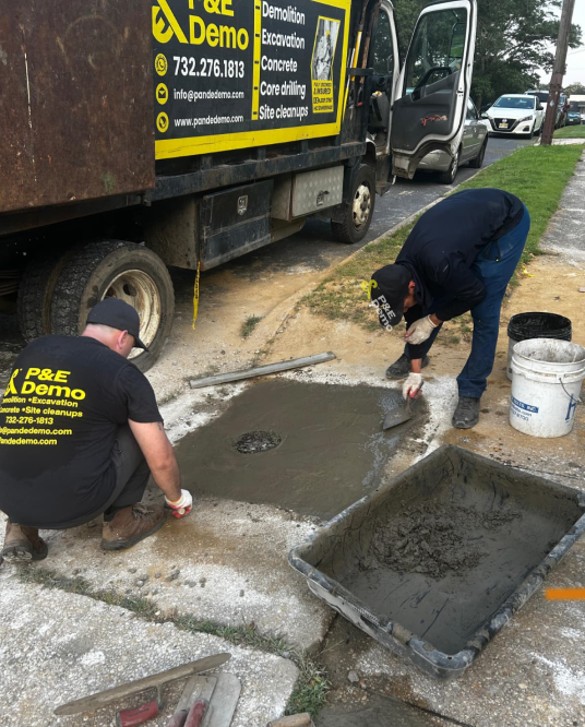 Two P&E Demo workers resurfacing concrete, one smoothing with a trowel while the other prepares material beside a mixing tray and company truck.