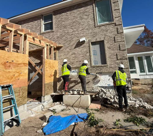 Three construction workers in safety vests and hard hats working on a brick house, with wooden framework, tools, and materials scattered around the site