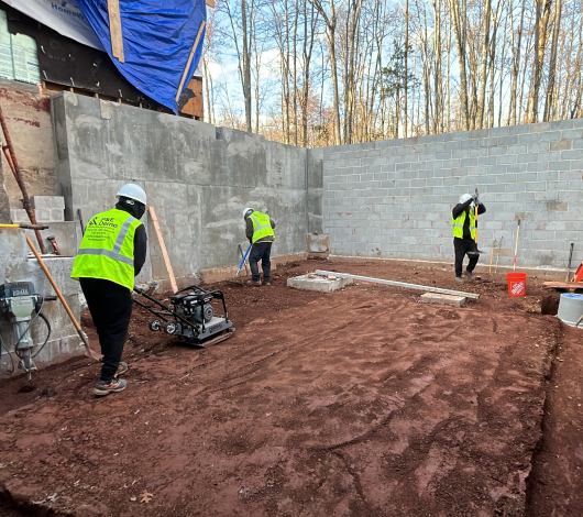 Three construction workers in safety vests and helmets preparing the ground inside a partially built structure, using a plate compactor, shovel, and rake