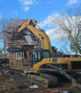 Yellow excavator tearing down a wooden house structure, representing P&E Demolition’s expertise in safe and controlled demolition.