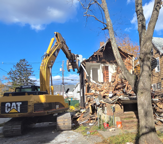Yellow Caterpillar excavator demolishing the front of a two‑story house in a residential neighborhood, with debris scattered and adjacent homes visible under a bright blue sky.