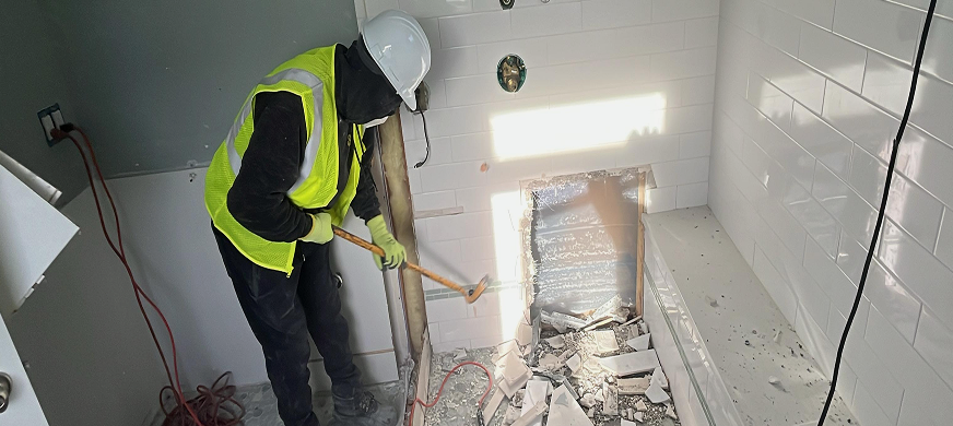 Construction worker in safety gear using a sledgehammer to demolish tiled bathroom wall during professional interior demolition in New Jersey