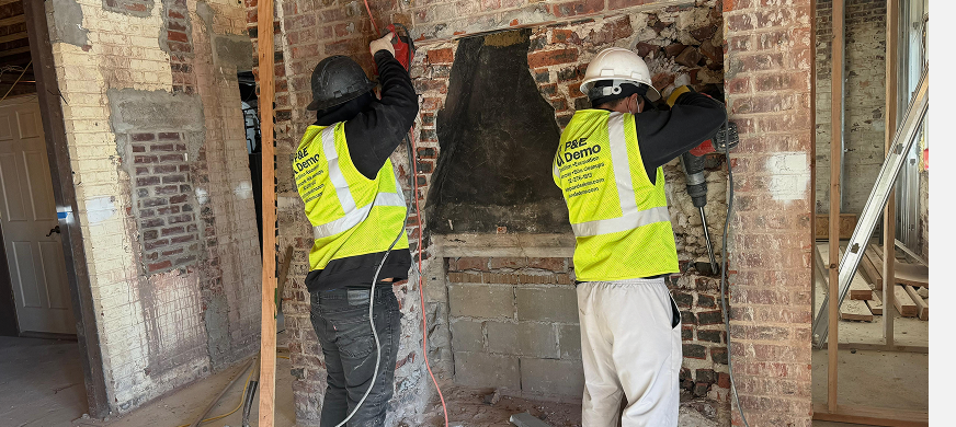Two demolition contractors in safety gear dismantling a brick chimney wall with power tools during professional chimney removal in New Jersey