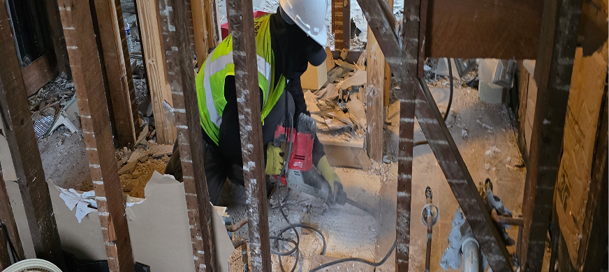 NJ demolition worker using a jackhammer to break up flooring during an interior demolition project, surrounded by exposed studs and construction debris
